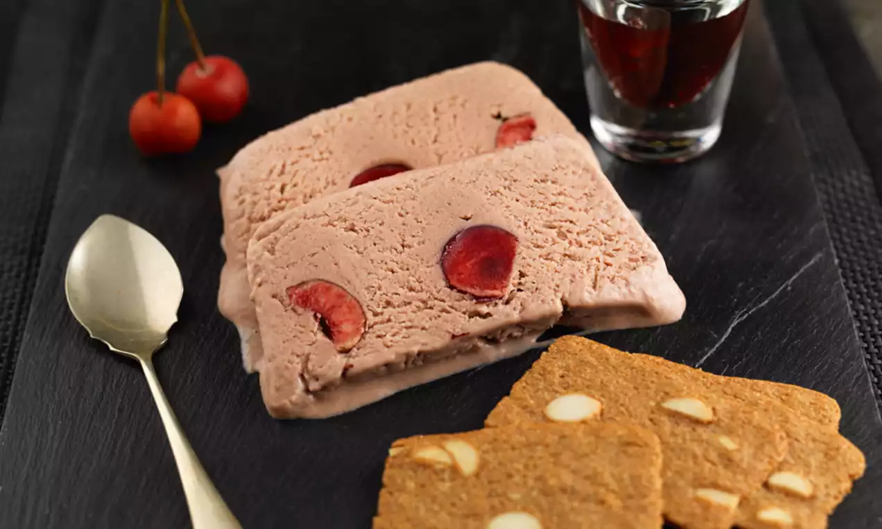 Cherry semi-freddo with rich pink creamy slices dotted with cherry halves on a dark slate plate alongside almond biscuits and fresh cherries