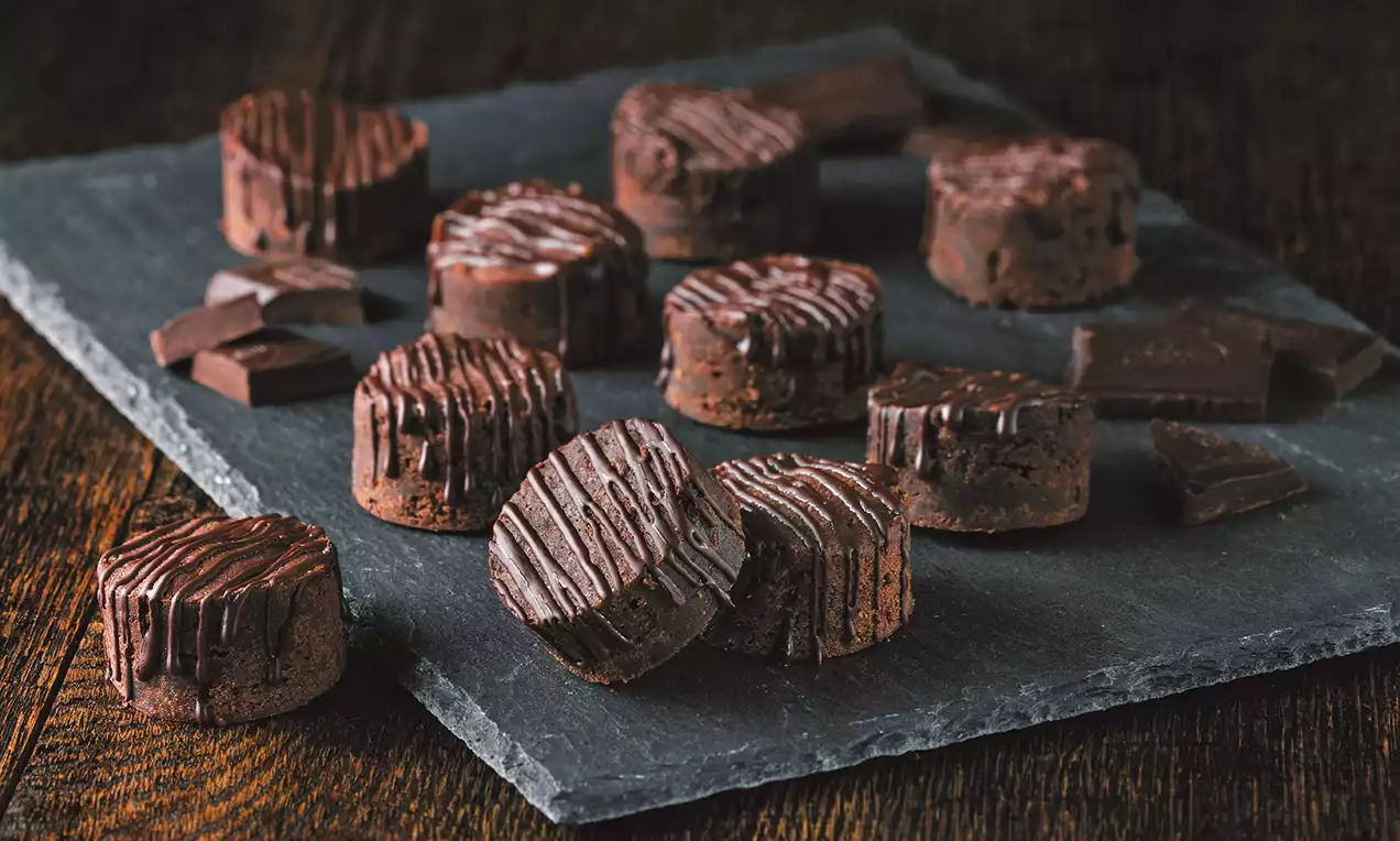 Kleine ronde chocoladekoekjes met donkerbruine geglazuurde strepen, gerangschikt op een donkergrijze steen, met stukjes pure chocolade eromheen