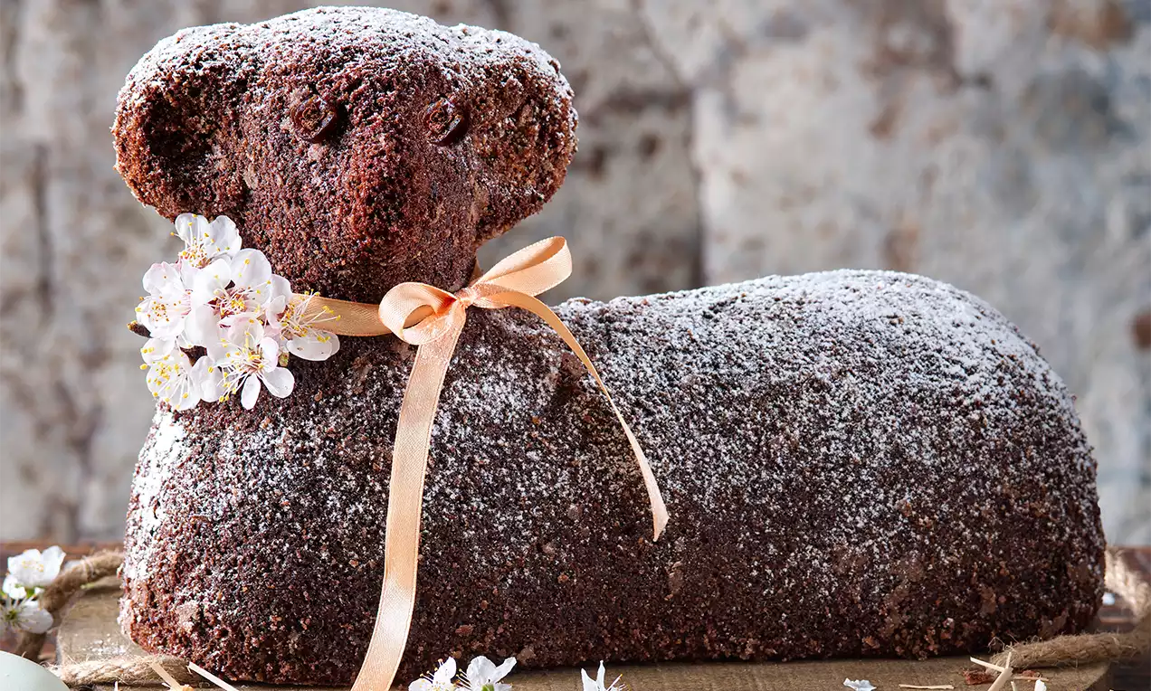 Tmavě hnědý kakaový beránek s jemnou strukturovanou polevou, posypaný moučkovým cukrem, ozdobený stuhou a kvítky na přírodním podkladu