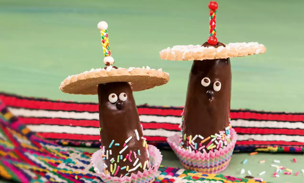 Schokoladenüberzogene Bananen in bunten Papierförmchen mit Zuckerdekor, kleinen Augen und Keks-Hüten auf gemustertem Stoff drapiert