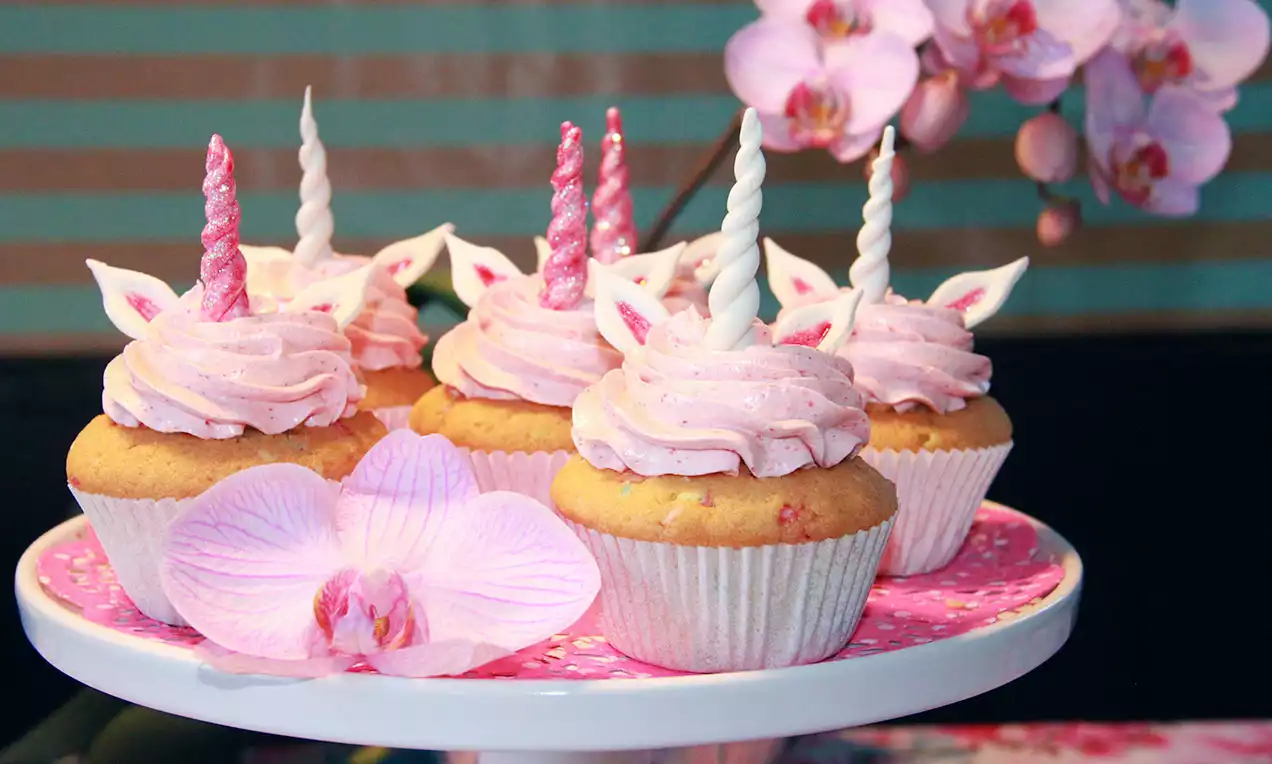 Einhorn-Cupcakes mit rosa Sahnetopping, glitzernden Hörnchen und Ohren auf weißer Platte mit pinker Orchidee und Streifenhintergrund