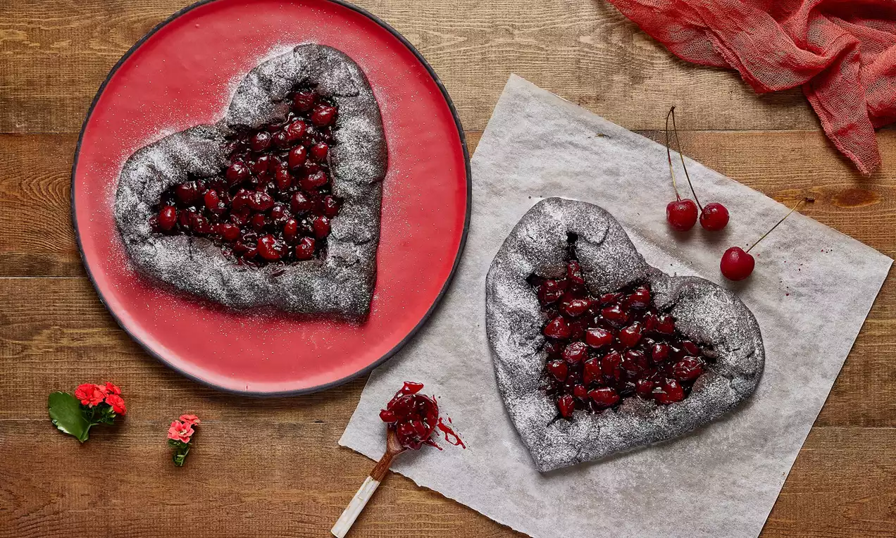 Kalp şeklinde, kenarları çatlak dokulu çikolatalı galette, parlak vişnelerle dolu, pudra şekeri serpilmiş ve yanına taze vişneler eklenmiş