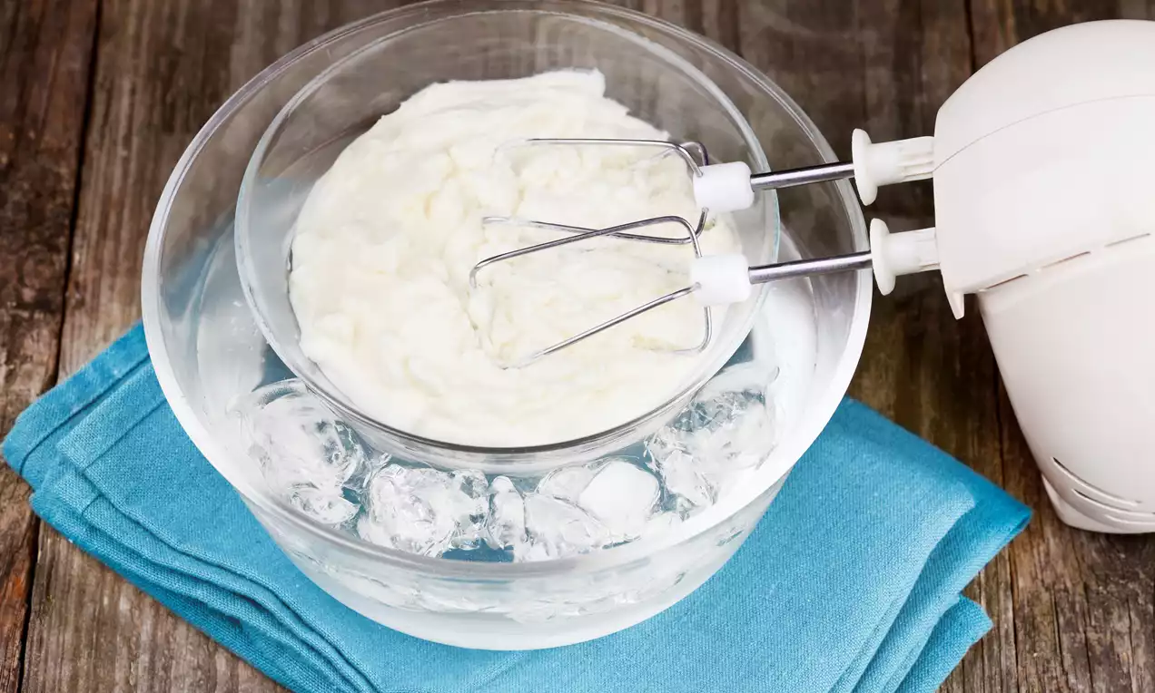 Śmietana kremówka ubijana metalowymi trzepaczkami w szklanej misce, ustawionej na lodzie, na niebieskiej ściereczce na drewnianym tle