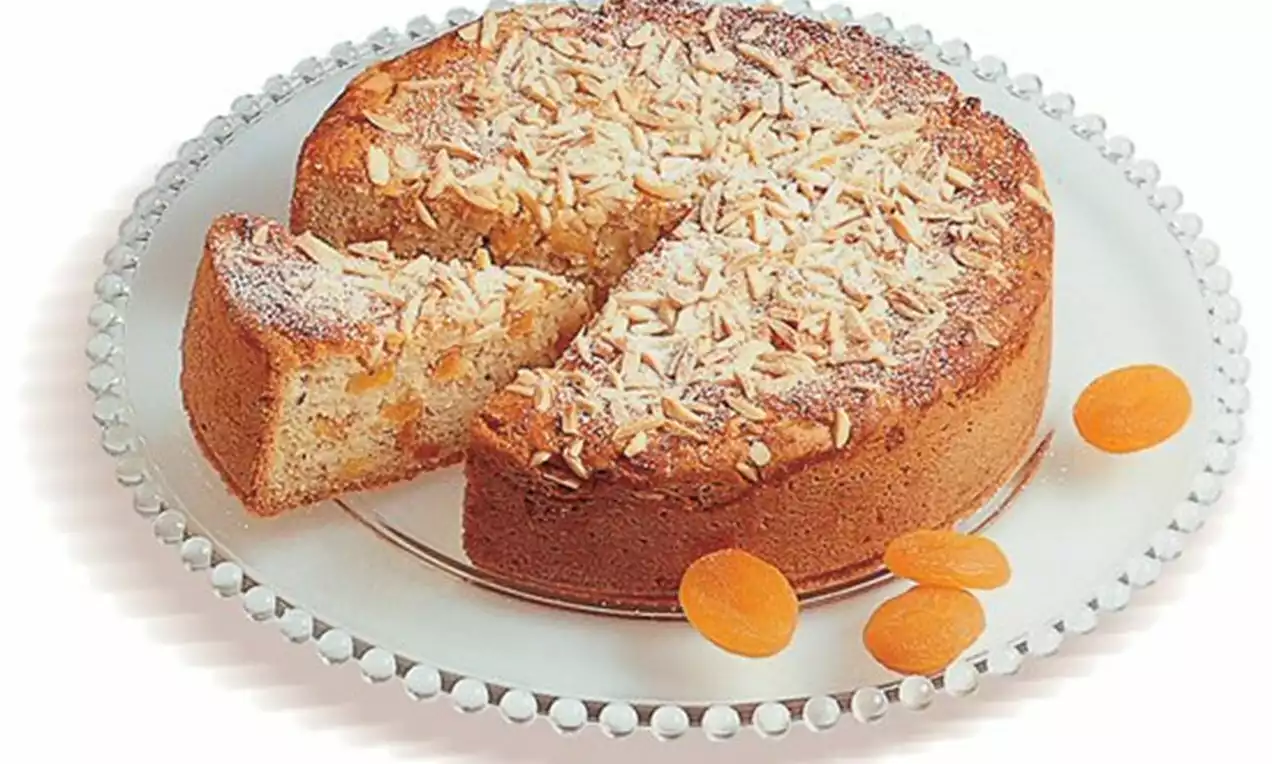 Plăcintă aurie cu mere și caise, de formă rotundă, acoperită cu fulgi de migdale pe o farfurie transparentă decorată subtil