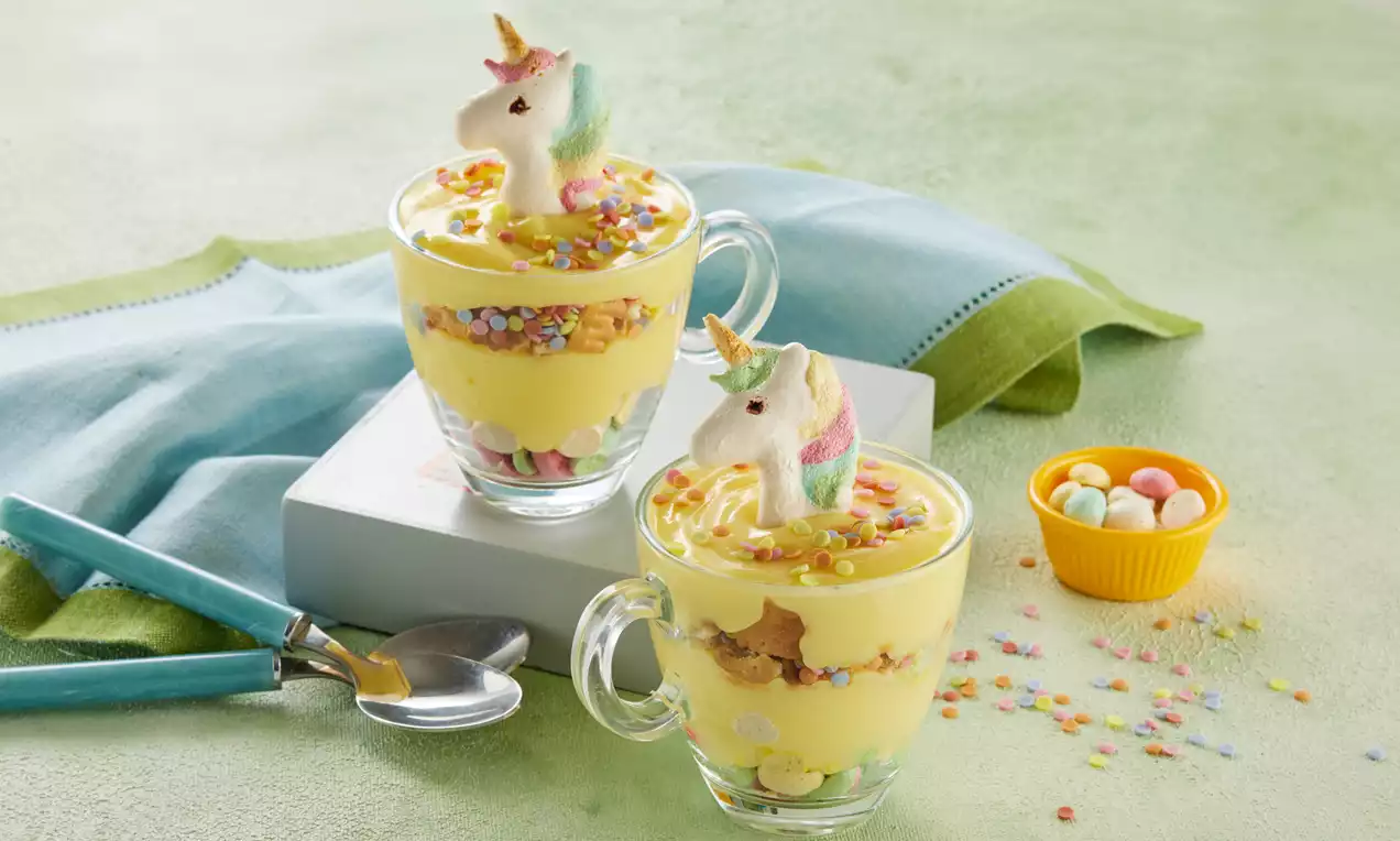 Cam bardaklarda sarı puding dolgusunun üzerinde renkli şekerlemeler ve unicorn figürü, yeşil masa örtüsü ve pastel tonlarda aksesuarlar eşliğinde
