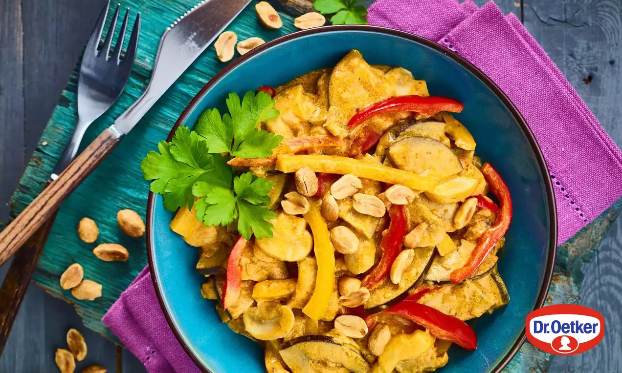 Veganské zeleninové kari v modré misce, zářivě žlutá omáčka s kousky červené papriky, cukety a arašídů, zdobené snítkou petržele na fialovém prostírání