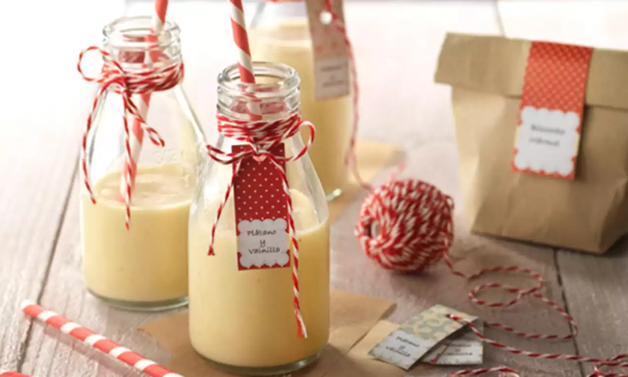 Smoothies cremosos color amarillo pálido en botellas de vidrio decoradas con etiquetas y lazos rojos, con pajitas rayadas sobre una mesa de madera