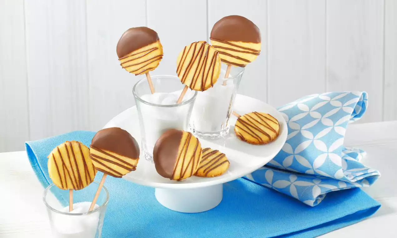 Runde Kekse mit gelber Oberfläche und braunen Streifen, teils mit Schokoladenglasur, auf Holzstäben, arrangiert auf weißem Teller mit blauer Serviette
