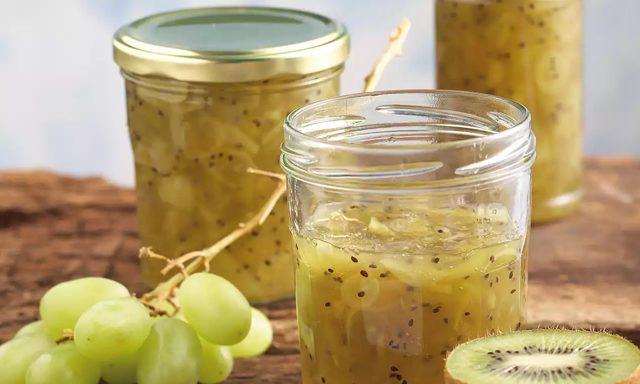 Confiture jaune translucide avec grains noirs, remplissant des pots en verre sur une table en bois, entourée de raisins verts et de kiwi en tranches