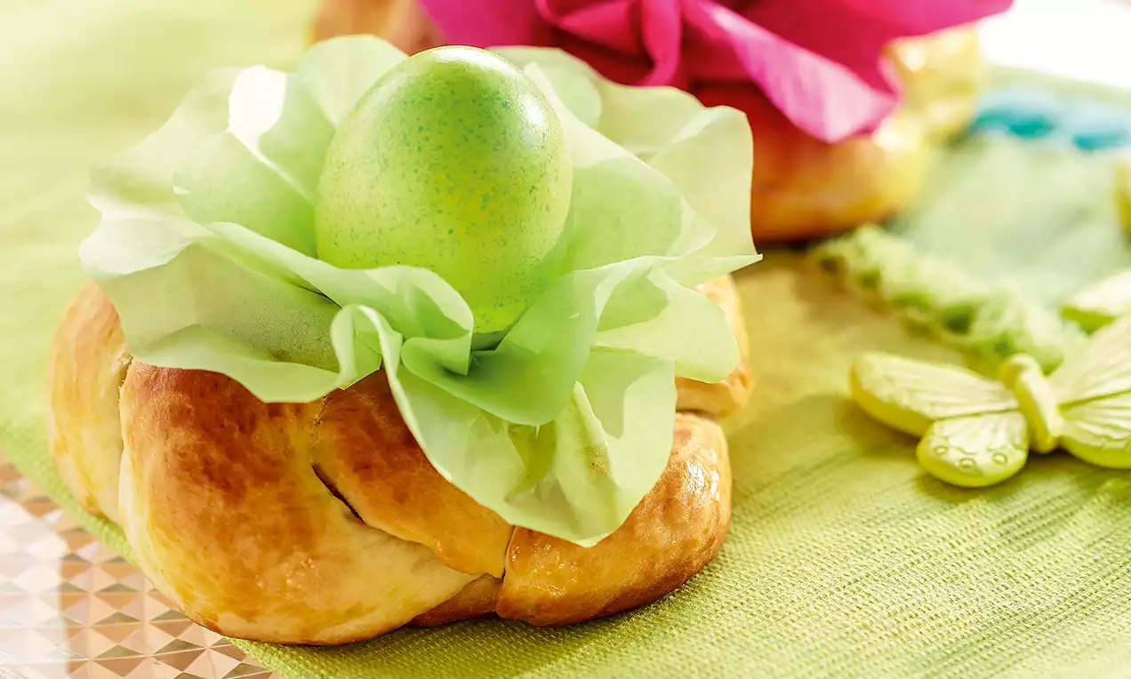Petite couronne briochée dorée garnie d'un œuf vert brillant enveloppé de papier vert pâle, posée sur une nappe printanière
