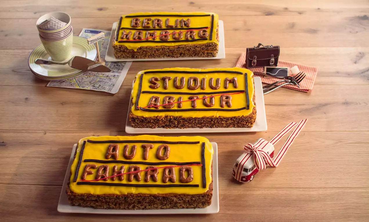 Rechteckige Kuchen mit gelbem Zuckerguss und roter Schrift auf weißer Platte, umgeben von Accessoires wie Tasse, Kamera und Modellauto