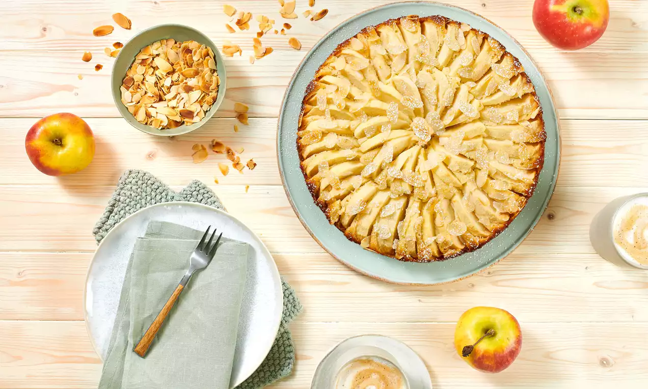 Goldbrauner Apfel-Marzipan-Kuchen mit dekorativ angeordneten Apfelspalten auf grünem Teller, umgeben von Äpfeln, Mandelblättern und Geschirr