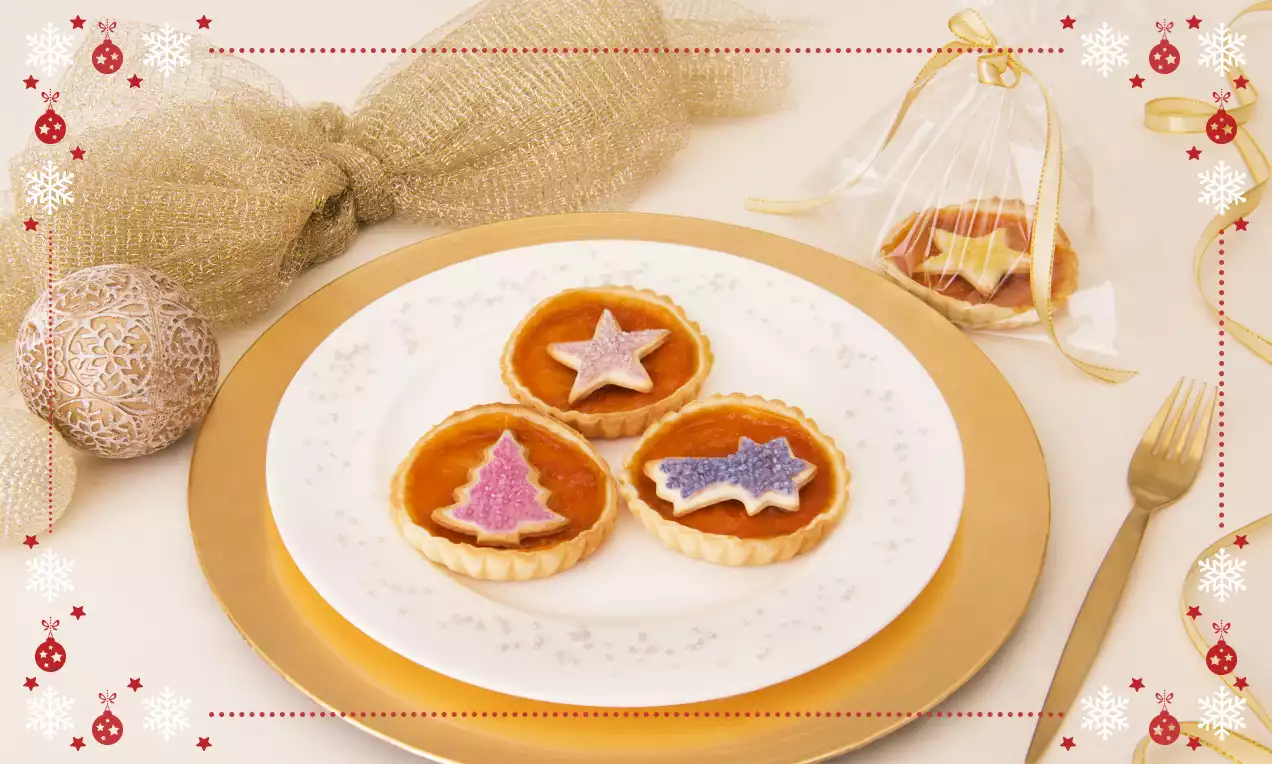 Crostatine tonde con base dorata e ripieno ambrato, decorate con biscotti di Natale glassati a forma di stella, albero e cometa su un piatto bianco decorato