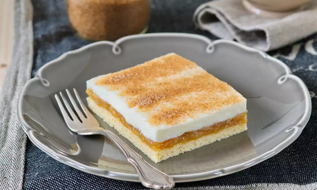 Světlý hranatý zákusek na šedém talíři, vrstvy světlého těsta, hnědé jablečné náplně a bílé polevy posypané skořicí vedle stříbrné vidličky