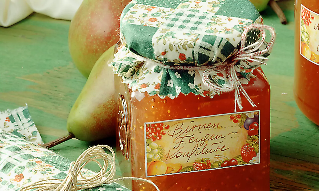 Ein viereckiges Glas mit orangefarbener Birnen-Feigen-Konfitüre, verziert mit kariertem Stoffdeckel und Schleife, vor reifen Birnen drapiert