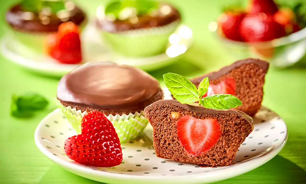 Wilgotne babeczki czekoladowe z błyszczącą polewą, truskawkowym wnętrzem w kształcie serca, na białym talerzu w kropki z listkiem mięty