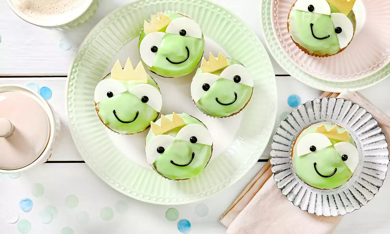 Froschkönig-Muffins mit grüner Glasur, goldenen Kronen und schwarzen Zuckerdekor-Augen auf pastellfarbenen Tellern und weißem Tisch drapiert