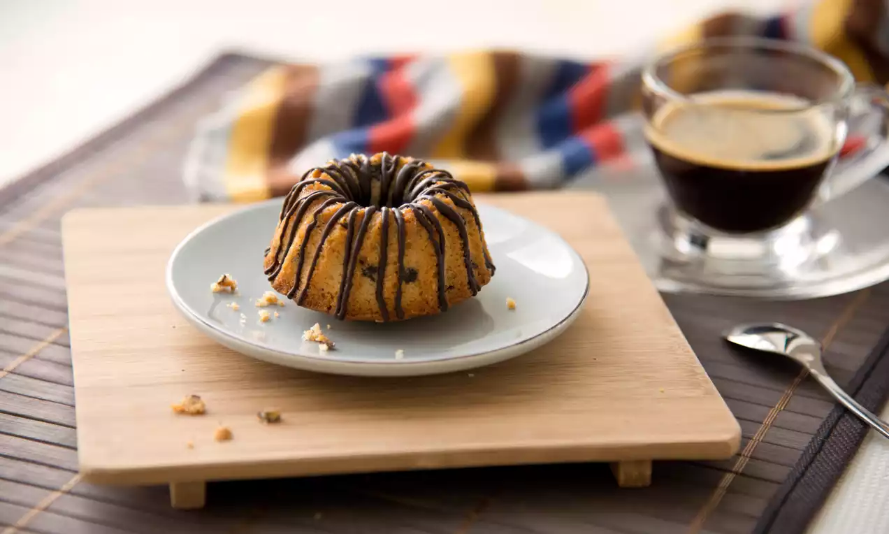 Kleiner, goldbraun gebackener Gugelhupf mit Schokostückchen und dunkler Glasur auf einem hellblauen Teller auf Holzbrett serviert