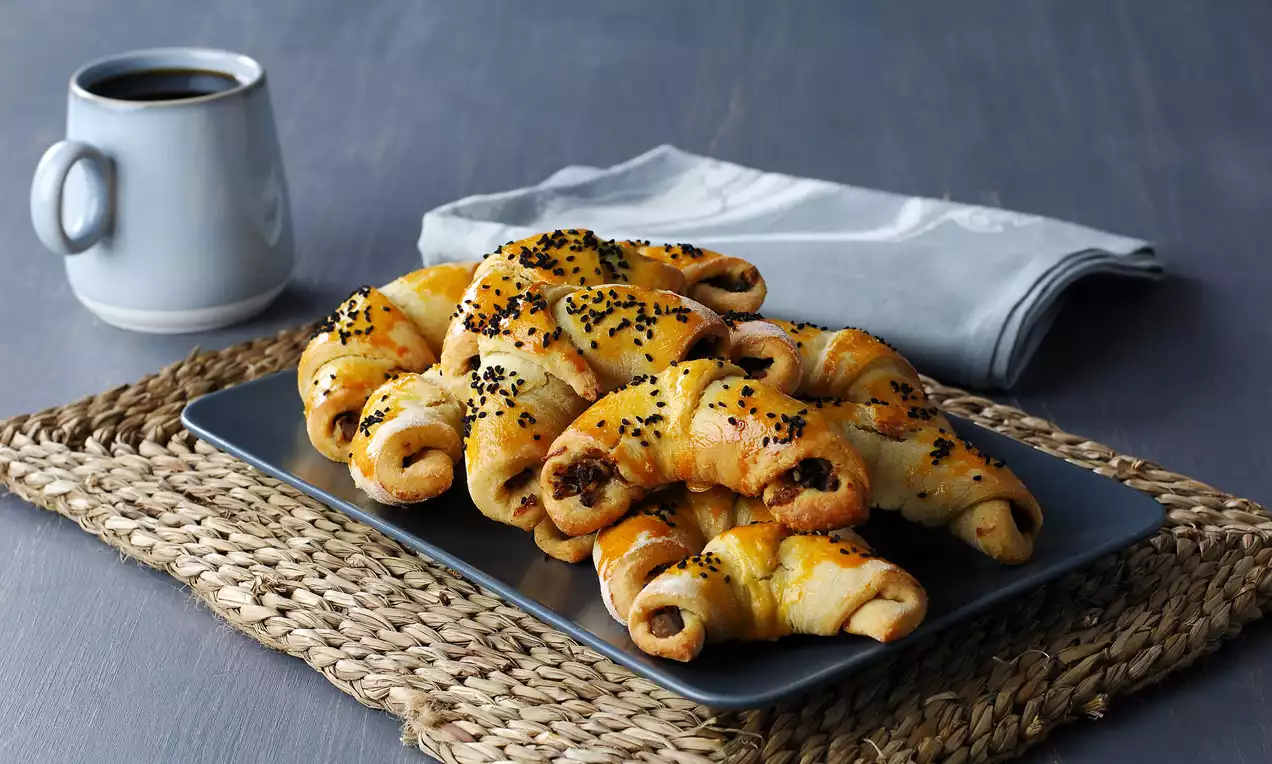 Parlak altın rengi hamur, içi patlıcan harcıyla dolu, kıvrık şekilli ay çörekleri siyah susamla süslenmiş tabağa dizili