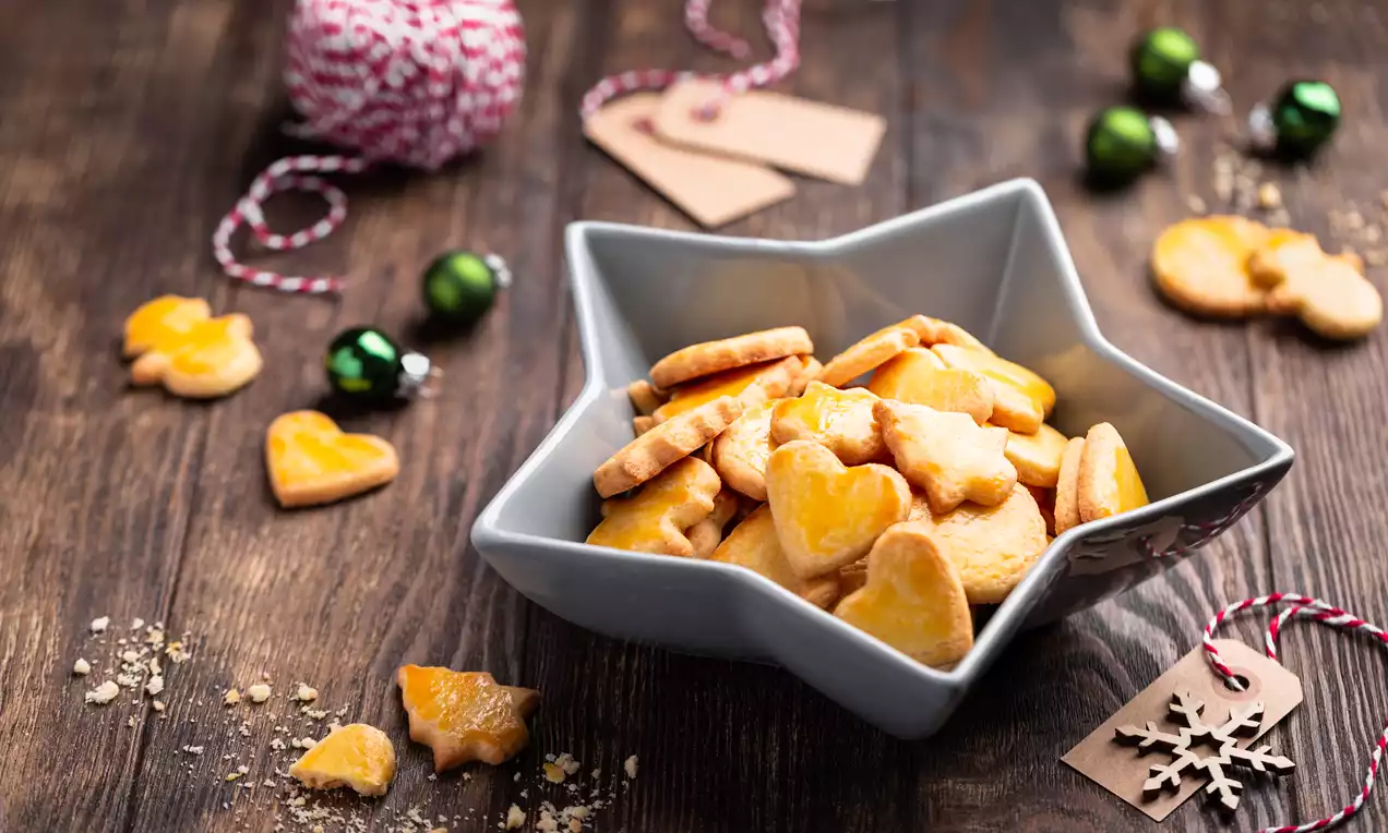 Goldgelbe, glänzende Mailänderli in Sternform und Herzform in einer grauen, sternförmigen Schale auf rustikalem Holztisch mit Weihnachtsdekoration