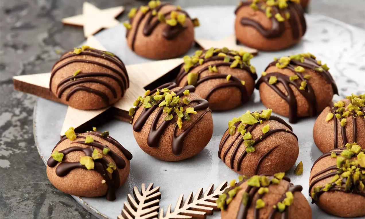 Runde, schokoladenbraune Kugeln mit glänzenden Schokolinien und gehackten Pistazien auf einer grauen Platte mit Holzstern und Zweigdeko