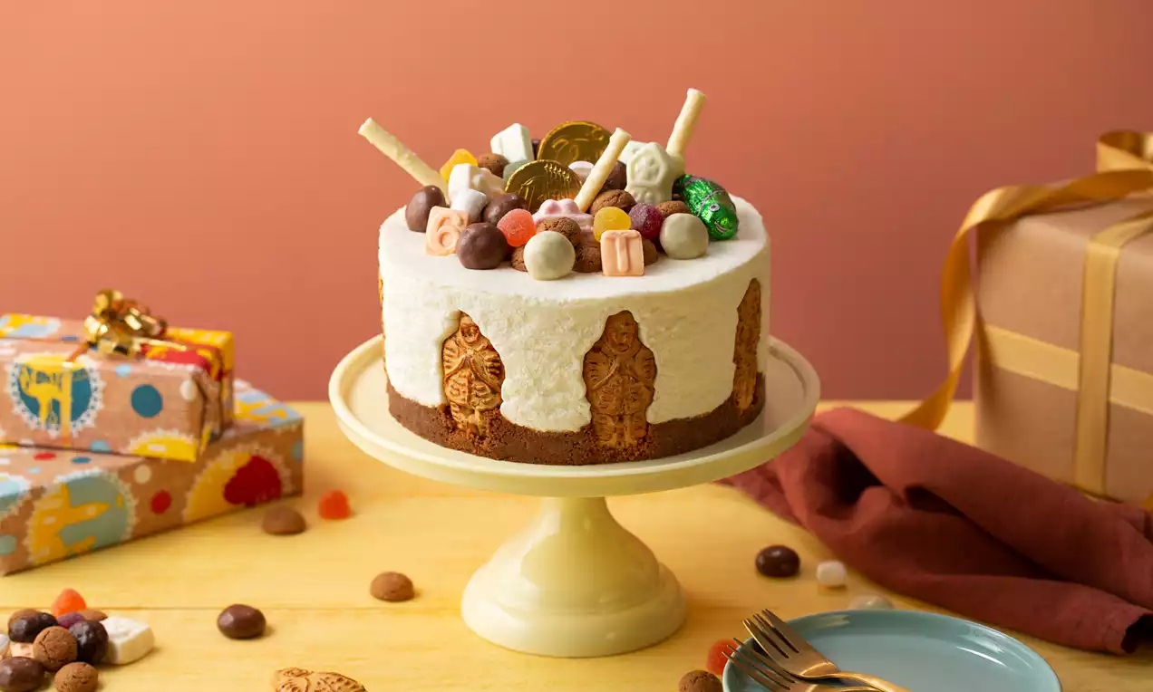 Sinterklaas MonChoutaart met witte roomlaag, bruine bodem, versierd met taaitaai, chocolade, marsepein en gouden munten op een lichtgele taartstandaard