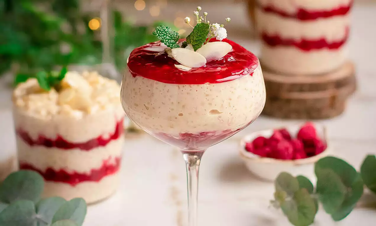 Svijetli kremasti sutlijaš poslužen u staklenoj čaši na stalku, preliven sjajnim crvenim prelivom od malina i ukrašen listovima mente