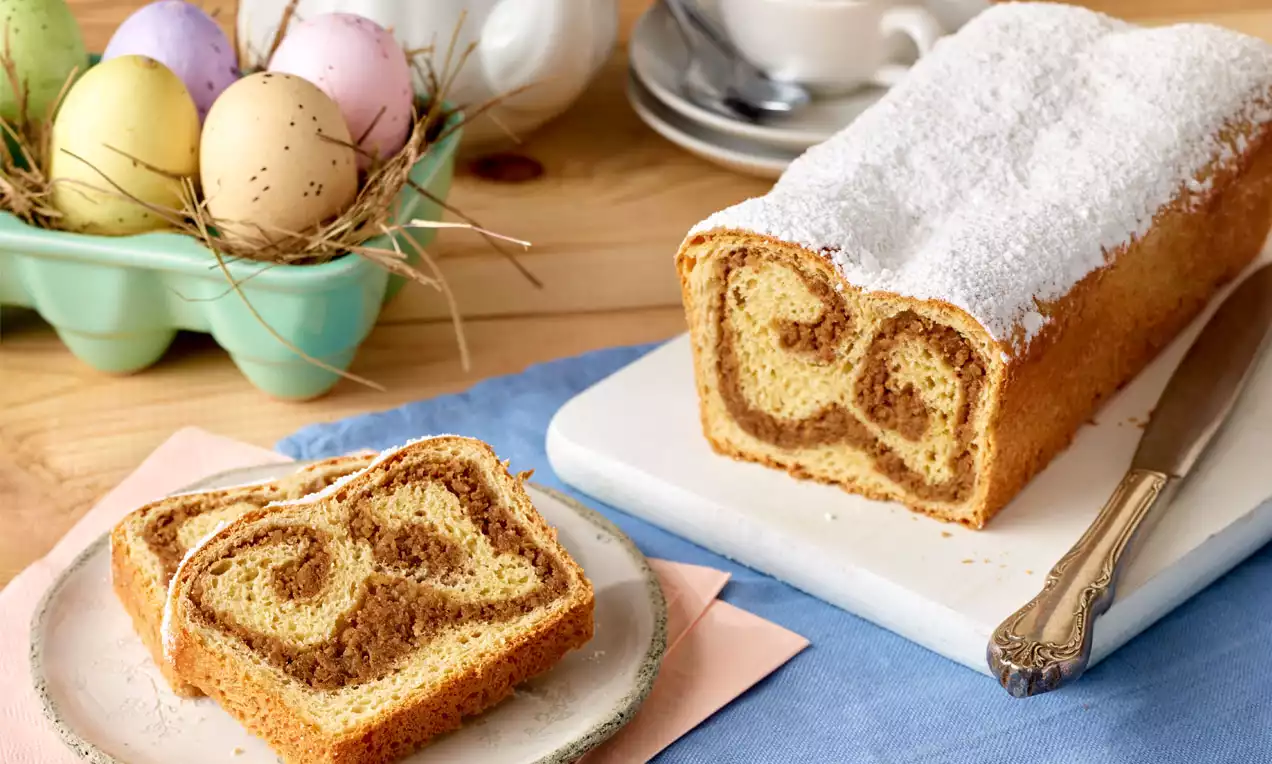 Hellbraune Nusspotitze mit weißem Zuckerguss, feiner, gewundener Füllung auf Holzbrett serviert, daneben eine Scheibe auf Teller