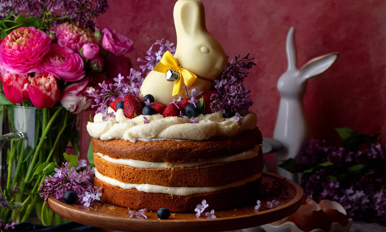 Торта с три златисто-кафяви блата, междинно кремообразно пълнеж, гарнирана с бели цветя, горски плодове и шоколадов заек върху дървена стойка