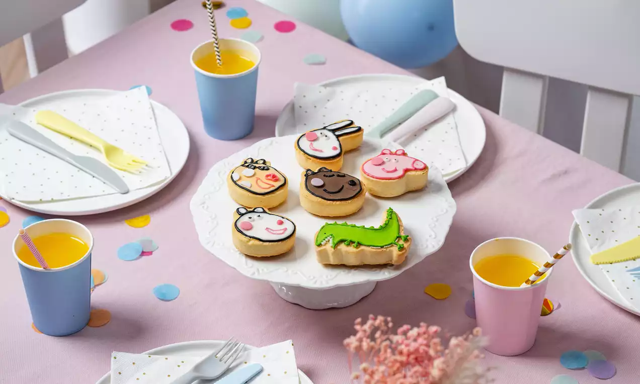 Bunte Peppa Wutz Küchlein mit glatter Zuckerglasur und gezeichneten Figuren auf weißer Tortenplatte, umgeben von farbiger Partydekoration