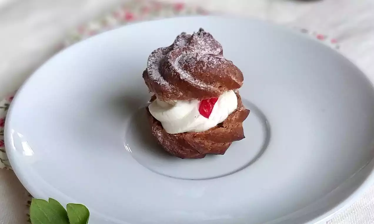 Kakaová profiterolka s vrstvami tmavě hnědého těsta, nadýchanou bílou šlehačkou, jasně červeným ovocem a jemným popraškem cukru na bílém talíři