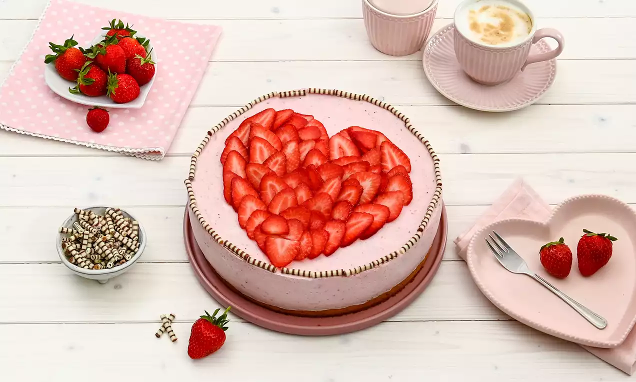 Gâteau rond rose clair garni de tranches de fraises disposées en forme de cœur, avec perles chocolatées sur une assiette rose