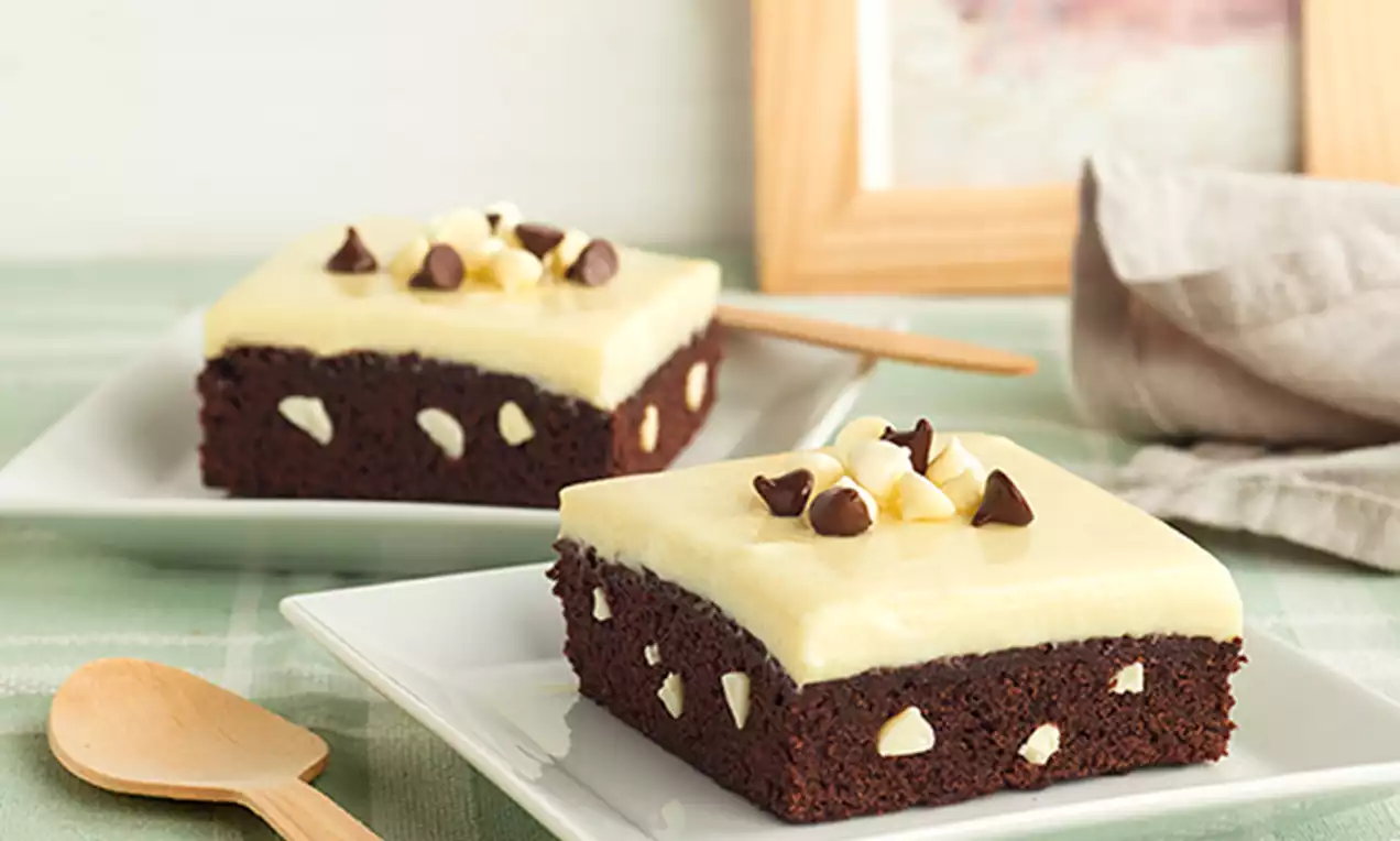 Cuadrados de bizcocho negro con chips blancos integrados y cobertura cremosa vainilla, decorados con gotas de chocolate en platos blancos