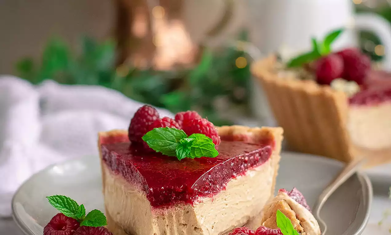 Tart trouglog oblika sa svijetlom kremom, crvenim premazom i malinama, ukrašen listićima mente na svijetlom tanjuru okružen zelenilom