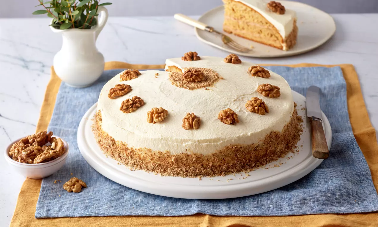 Helle Nusstorte mit Walnüssen dekoriert auf weißer Platte, umgeben von blau-gelbem Stoff, daneben Messer und kleine Walnussschale