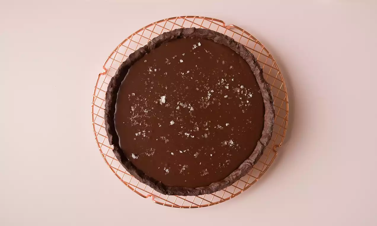 Tarta redonda de chocolate oscuro con borde crujiente, superficie lisa salpicada de cristales de sal, colocada sobre una rejilla metálica