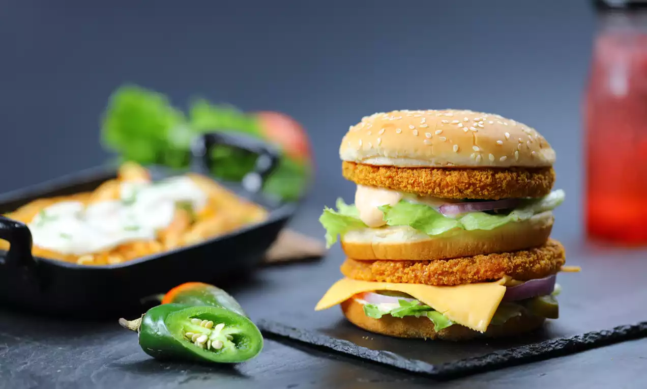 Golden sesame bun layered with crisp breaded patty, leafy lettuce, melted cheese, onion and creamy sauce on a slate with vibrant sides