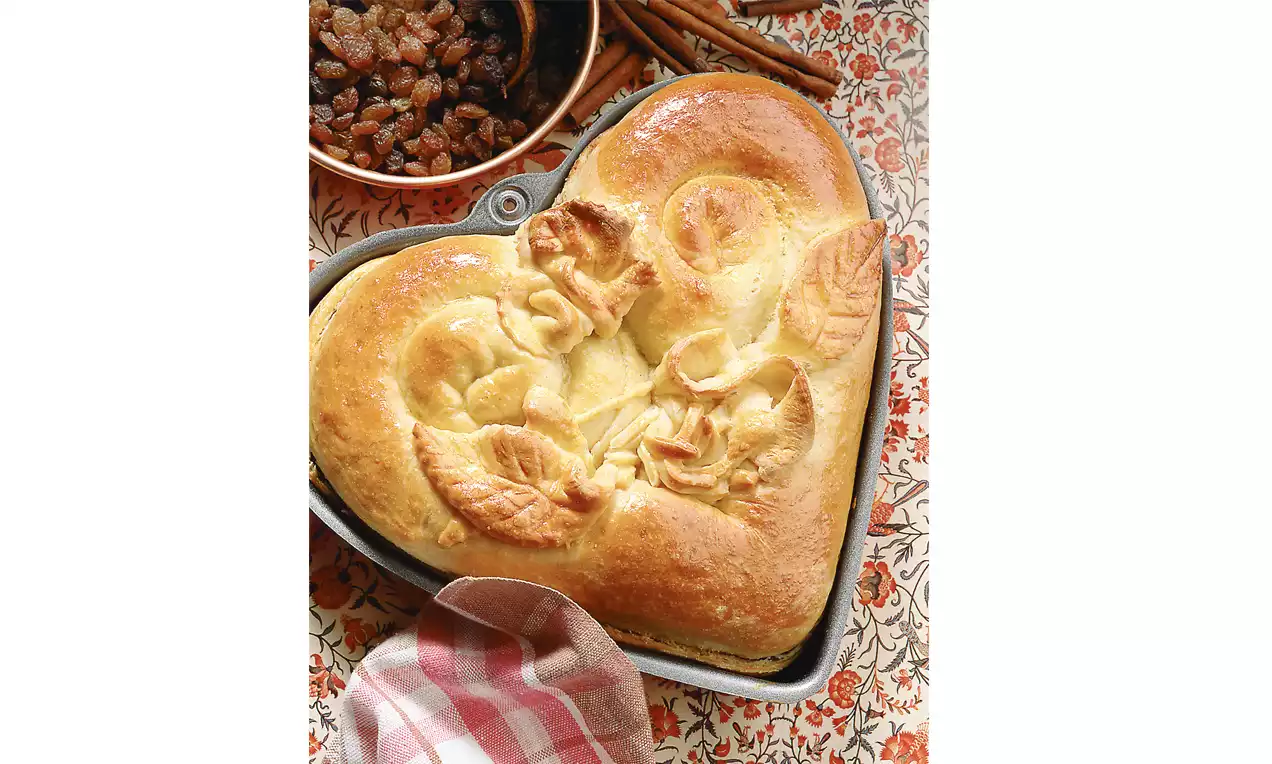 Pain doré en forme de cœur avec reliefs feuilles et spirales dans un moule gris, posé sur un tissu à motifs rouges avec raisins secs et bâtons de cannelle