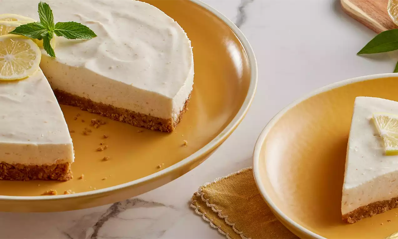 Kremalı beyaz yüzeyi ve kıtır bisküvi tabanı olan cheesecake, limon dilimleri ve nane yaprağı ile altın sarısı tabakta sunulmuş