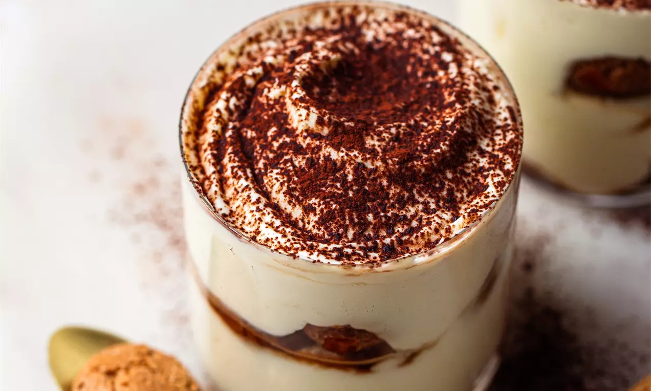 Picture - Layered almond tiramisu in clear glasses with pale cream filling, espresso-soaked biscuits, cocoa dusting, gold spoon and amaretti