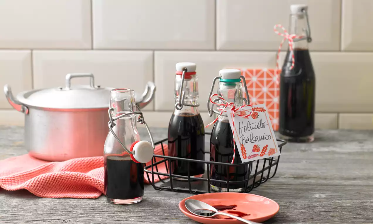 Dunkelbraune Flüssigkeit in schlanken Glasflaschen mit Verschluss in schwarzem Drahtkorb, daneben rosafarbene Stoffserviette und Teller