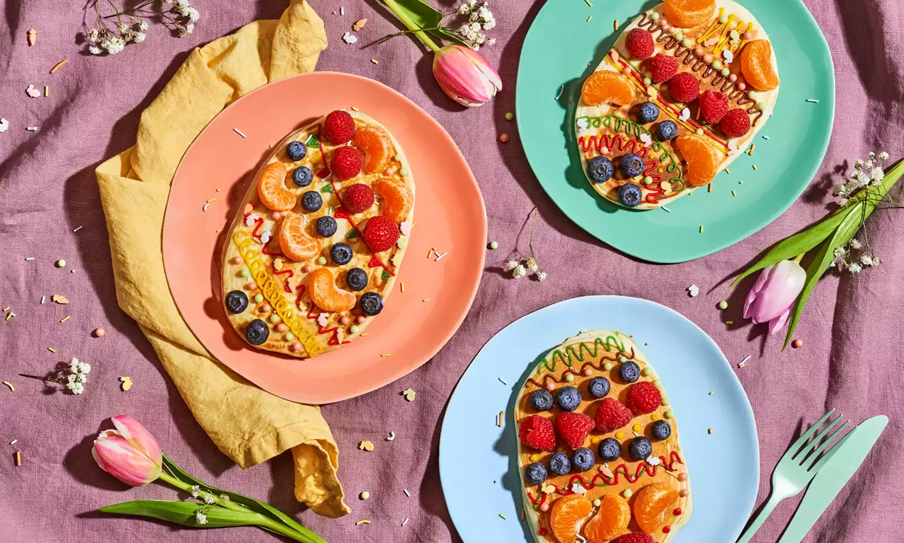 Ovale American pancakes met kleurrijk fruit zoals blauwe bessen, mandarijn en framboos, versierd met strepen glazuur op pastel borden met bloemen