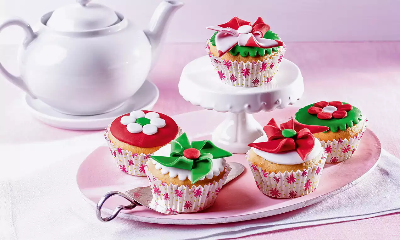 Petits muffins dorés ornés de fleurs rouges et vertes en pâte à sucre, disposés sur un plateau rose à côté d'une théière blanche