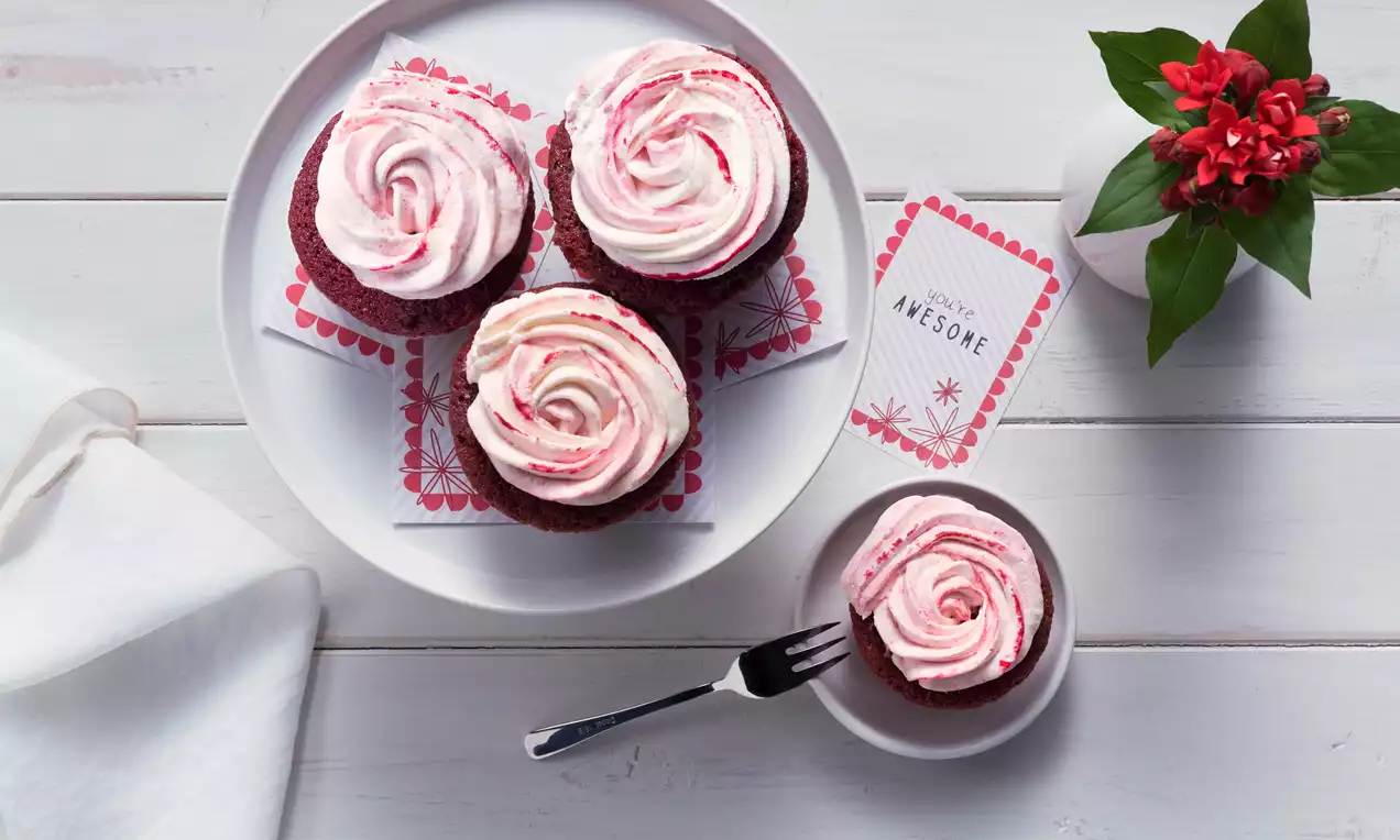 Rote Red Velvet Cupcakes mit zartrosa Cremehauben in runden Formen auf weißem Teller mit dekorativen Karten und rotem Blütenzweig