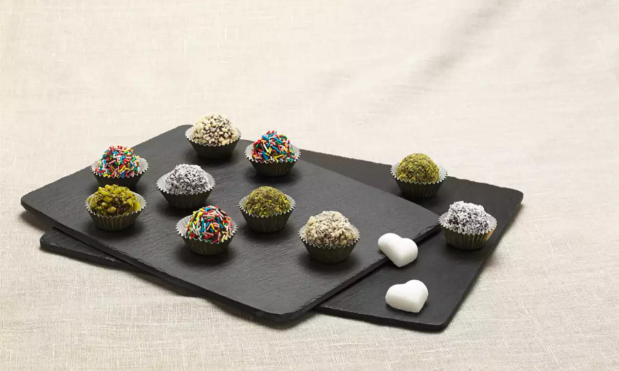 Tartufi rotondi colorati con granella di pistacchio, zucchero a velo e codette, disposti su piatti di ardesia nera accanto a due cuoricini bianchi
