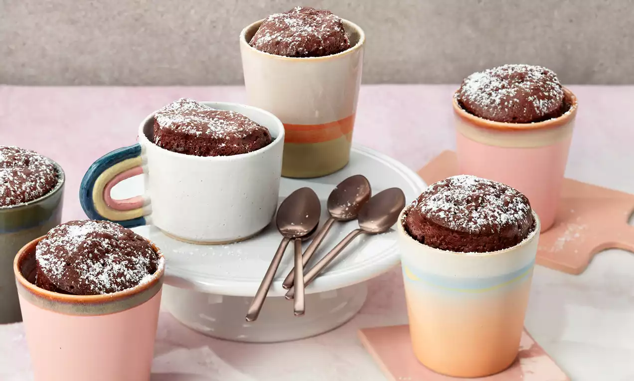 Schokoladige Tassenkuchen mit staubzuckerbedeckten, aufgewölbten Oberflächen in bunten Keramiktassen, angerichtet mit Löffeln auf einer weißen Tortenplatte