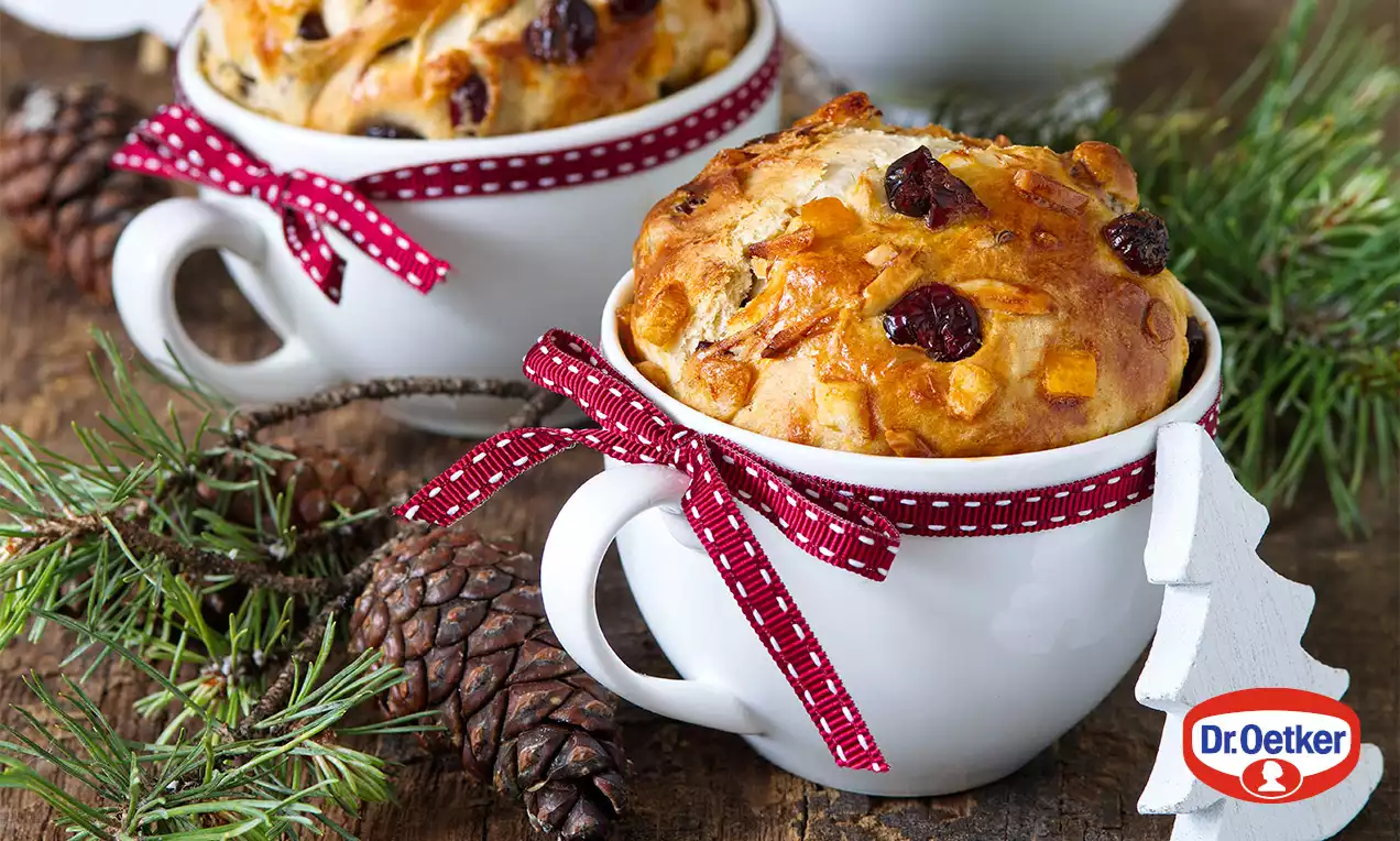 Zlatohnědé brusinkové dezerty s křupavou kůrkou v bílých hrníčcích, ovázané červenou stuhou, na dřevěném stole s větvičkami a šiškami