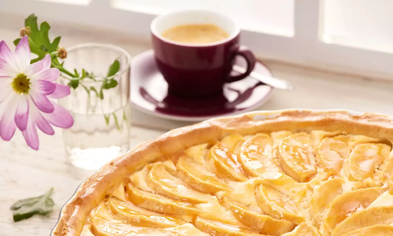 Tarte aux pommes dorée avec des tranches fines disposées en cercle, bord légèrement croustillant, accompagnée d'une tasse de café et d'une fleur