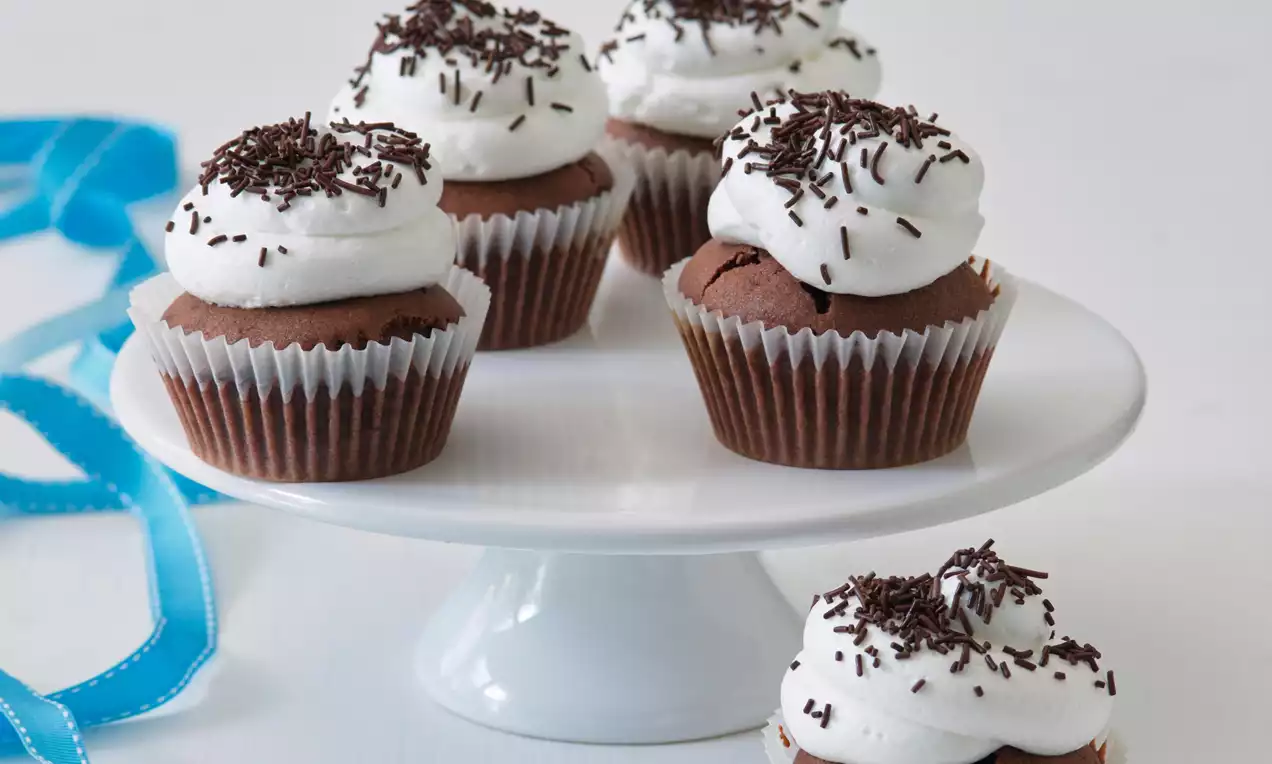 Čokoládové cupcakes s nadýchaným bílým krémem, dekorované tmavými čokoládovými hoblinkami na bílé stojánkové míse s modrou stuhou