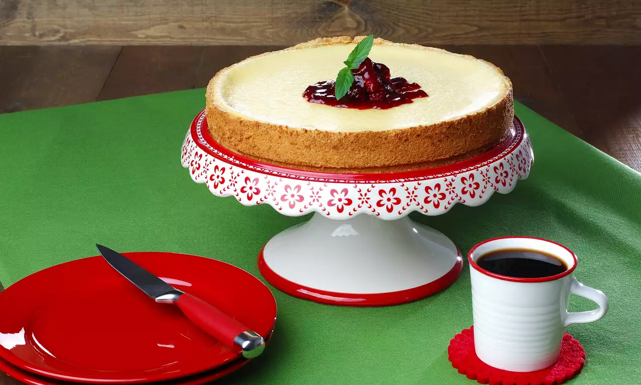 Altın rengi tabanlı yuvarlak cheesecake, üzeri vişne sosu ve nane yaprağıyla süslenmiş, kırmızı detaylı beyaz servis tabağında
